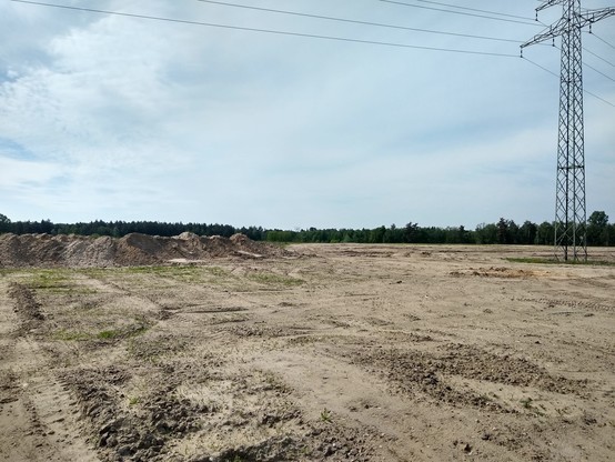 A flat wide landscape filled with sand. There are a few piles of sand far to the left. A high voltage power line is crossing left to right, with a pylon to the right. A forest can be seen far on the horizon.