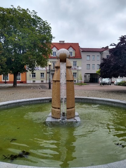 Okrągła fontanna pośrodku placu. Pośrodku fontanny znajduje się kamienny kwadratowy blok w kolorze popielatym. Na nim stoją cztery kamienne podpory w kolorze brązowym, na których szczycie oparta jest kamienna kula tej samej barwy. Podpory mają przekrój wycinków okręgu. Zwężają się ku górze. Przy spodzie nacięte są wąskim, poziomym wgłębieniem. Przy górze z kolei dwukrotnie następuje poszerzenie na kształt "kapturka".

Pośrodku kwadratowego bloku znajduje się dysza, która wystrzeliwuje wodę w gó…