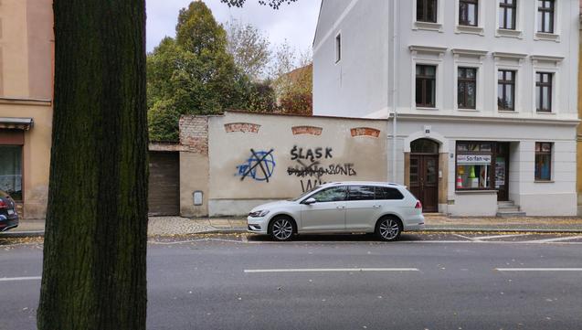 Fragment ulicy w niemieckim mieście. Pośrodku, za zaparkowanym białym samochodem, na ścianie widać bazgroły po polsku: po lewej "A" w kółko niebieskim sprejem, następnie przekreślone czarnym sprejem. Obok tym samym czarnym sprejem "Śląsk. Dynamo Zone. WKS", przy czym "Dynamo" również jest przekreślone. Po prawej stronie sklep "Sorfan" z pachołkami w oknie.