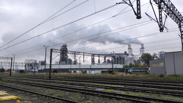 Obszar przemysłowy widziany przez tory kolejowe. Znajduje się na nim wiele wysokich kominów o różnych kształtach, z których unoszą się szare kłęby pary, które wiatr znosi w lewo. To wszystko na tle srebrzystego, zachmurzonego nieba.