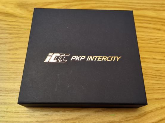 Ozdobny, prostopadłościenny granatowy kartonik na drewnianej półce. Na środku logo "PKP Intercity" złotymi literkami.