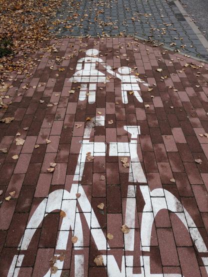 Chodnik z czerwoną kostką, na której namalowano dwa symbole: u góry osoba w szerokiej spódnicy, trzymająca dziecko za rękę, u dołu rower.