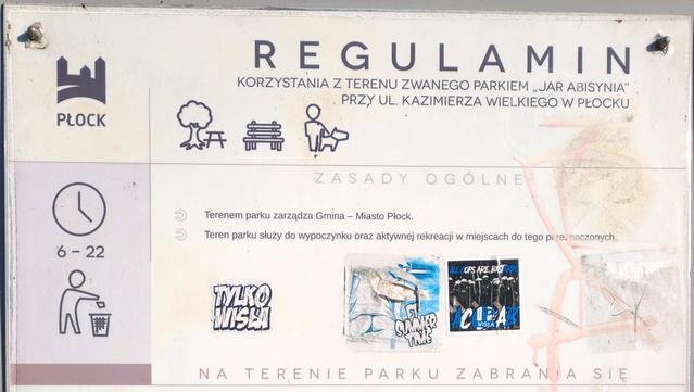 Tablica "Regulamin korzystania z terenu zwanego parkiem "Jar Abisynia" przy ul. Kazimierza Wielkiego w Płocku". Po lewej symbol miasta z napisem Płock, symbol zegara z godzinami 6–22 i człowieka wrzucającego śmieci do kosza. U góry symbole przedstawiające kolejno: stół przy drzewie, ławkę oraz człowieka z psem. Poniżej poszczególne zasady, w których już używa się krótkiego określenia "park". Na pustej przestrzeni pomiędzy zasadami ogólnymi i szczegółowymi nalepki lokalnego klubu sportowego i ch…