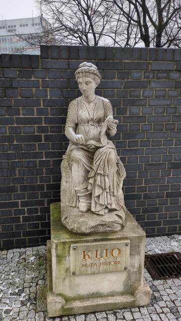 Pomnik na cokole, opisany "Klio, muza historii". Przedstawia siedzącą kobietę w sukni, z papirusem rozwiniętym w jednej dłoni, opadającym na kolana. Na głowie widać pionowe smugi spływającego deszczu, które nadają twarzy wyraz płaczu.