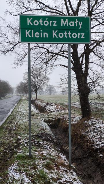 Zielona, przydrożna tablica z nazwą miejscowości. Na górze "Kotórz Mały", poniżej "Klein Kottorz". W tle rów, trochę śniegu na trawie, pojedyncze drzewa przy drodze.