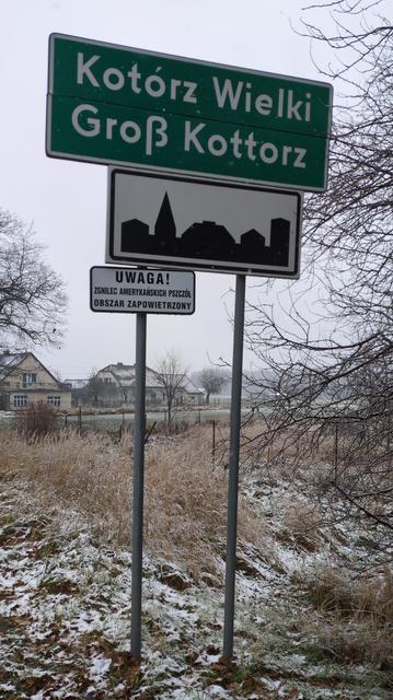 Podobna tablica, tym razem "Kotórz Wielki" i "Groß Kottorz". Poniżej znak początku obszaru zabudowanego i ostrzeżenie o zgnilcu amerykańskim pszczół. W tle lekko oprószone śniegiem żółte trawy, dalej domy.