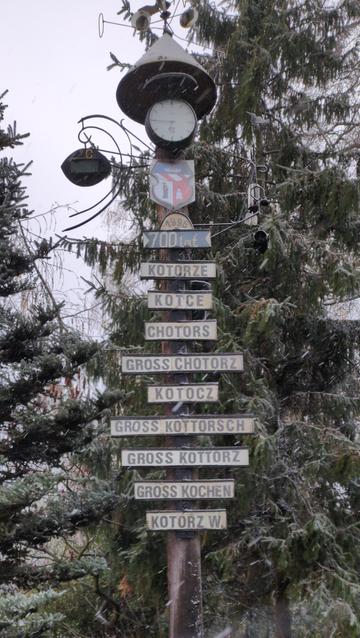 Słup ze stożkowatym daszkiem. Na górze zegar, herb i belka z napisem "700 lat" i rokiem 1995. Poniżej belki z kolejnymi nazwami: Kotorze, Kotce, Chotors, Gross Chotorz, Kotocz, Gross Kottorz, Gross Kochen, Kotórz W.