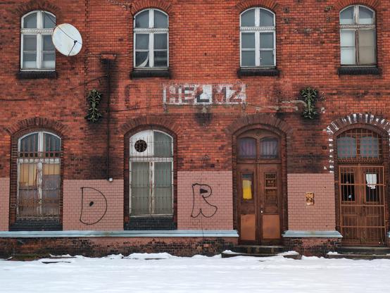 Budynek starego dworca kolejowego z czerwonej cegły. Pośrodku widać pozostały fragment starego napisu na białym tle "HEŁMZ". Nań nakłada się cień większego, prawie całkiem zmytego napisu "CHEŁMŻA", z którego tak naprawdę widać głównie "CH" na lewo od tego białego.