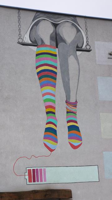 Mural przedstawiający nogi pindy, siedzącej na huśtawce, z kolorowymi rajstopami, na tle ściany koloru piaskowego. Wszystko poza rajstopami wykonane jest w odcieniach szarości. Widoczną część nóg wykonano szerokimi, jednobarwnymi pociągnięciami, łącząc szarą farbę z tłem ściany; część podudzia i kolana mają kolor tła. W odcieniach szarości wykonano również cienką, drewnianą huśtawkę z metalowymi łańcuchami, oraz spód szarej, krótkiej sukienki z białym wykończeniem. Rajstopy z kolei przedstawiaj…