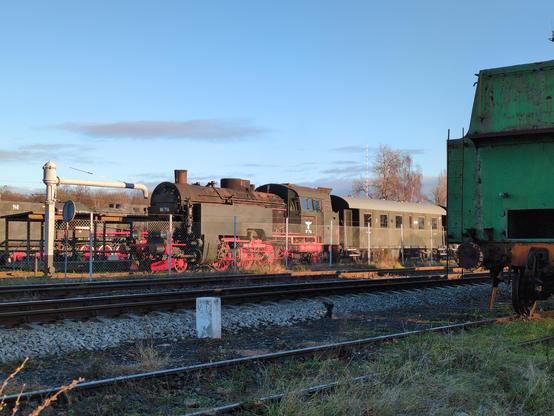 Lokomotywa parowa za płotem. Okrągłą część z kotłem ma czarną, z boku dostaje do niej oliwkowy, nitowany sześcian. Oliwkowa jest również kabina maszynisty. Koła i inne elementy podwozia są czerwone. Na czole ma numery 86 714. Na boku kabiny maszynisty ten same numer, choć trochę starty, a nad nim orzeł Trzeciej Rzeszy. Za lokomotywą oliwkowy wagon z otwartymi drzwiami, oznaczony jako 2 klasa. Z tyłu inna oliwkowa lokomotywa. Po prawej stronie fragment innej, zielonej lokomotywy.