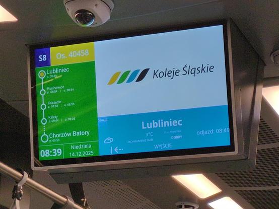Wyświetlacz w pociągu. W lewym górnym rogu "S8" na niebieskim tle i "Os. 40458" na żółtym. Poniżej rozkład jazdy, na zielonym tle: Lubliniec, trzy kolejne postoje i końcowy Chorzów Batory. Poniżej w tym samym kolorze czas 08:39 i data: Niedziela 14.12.2025.

Po prawej stronie duże białe polo z logotypem: cztery obłe cosie, trochę równoległoboczne, żółty, zielony, niebieski i czarny, i napis: Koleje Śląskie.

Poniżej, na niebieskim polu: stacja Lubliniec, piktogram chmurki, 3°C, zachmurzenie duż…