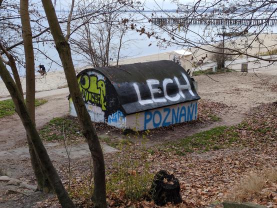 Budowla z zaokrąglonym dachem na brzegu plaży. Na dachu biały napis "LECH", poniżej na jasnej ścianie niebieski "POZNAŃ". Z tyłu plaża i morze, w oddali molo.