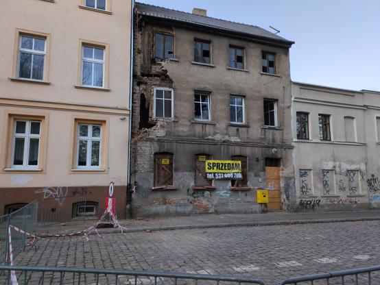 Trzypiętrowa, rozpadająca się kamienica, przyległa do kamienic w lepszym stanie. Na parterze okna i drzwi zabite są dechami, a na nich wisi napis "sprzedam" z numerem telefonu. Od pierwszego iętra wzwyż ściana frontowa odpadła z lewej strony, odsłaniając deski pod spodem i ziejącą, pustą przestrzeń, w której jakimś cudem trzyma się jeszcze okno. W oknach na piętrach szyby są częściowo powybijane, po prawej okno jest otwarte. Cała ulica wokół kamienicy ogrodzona jest płotkami ochronnymi, z biało…