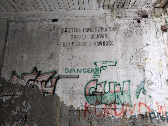 Ściana zrujnowanego budynku. U góry widać ślady równo wymalowanego napisu "Ojczyzna powierzyła tobie sprzęt bojowy — dbaj o jego sprawność." Poniżej bazgroły.