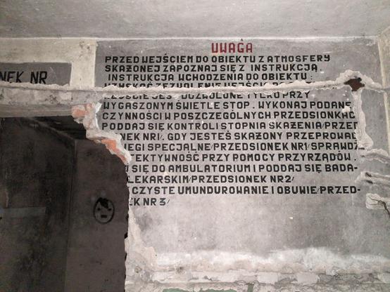 Częściowo uszkodzona instrukcja, namalowana na ścianie obok otworu drzwiowego. Część ściany po prawej stronie otworu została odłupana, uszkadzając napis. U góry został przerwany również, kiedy ze ściany wyrywano przewody.

W miarę możliwości rekonstruuję go do:
"""
Uwaga
Przed wejściem do obiektu z atmosfery skażonej zapoznaj się z instrukcją wchodzenia do obiektu: Uzyskać zezwolenie wejścia do [obiektu?]. Przejście jest dozwolone tylko przy wygaszonym świetle stop. Wykonaj podane czynności w p…