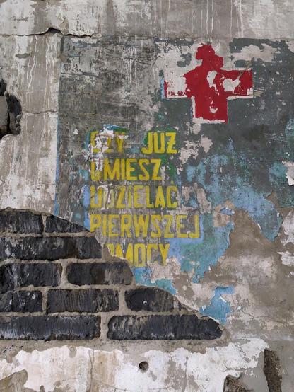 Fragment napisu na ścianie. Dół tynku oderwany, odsłaniając ciemne cegły. Żółtymi literami "czy już umiesz udzielać pierwszej pomocy". Nad napisem duży, czerwony krzyż.