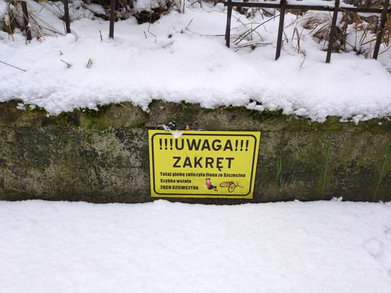 Żółta tabliczka na podmurówce. Na niej:

"""
!!!Uwaga!!!
Zakręt.
Tutaj glebę zaliczyła Ilona ze Szczecina.
Szybko wstała.
ZUCH DZIEWCZYNA.
"""

Na dole schematyczny rysunek przewróconego rowera, a obok niej siedzącej na czterech literach pindy w czerwonej koszulce z białymi rękawami, granatowych spodniach i jasnobrązowych butach. Ma krótkie, brązowe włosy z kitką z boku. Niestety, jej twarz jest całkiem gładka — nie ma oczu, nosa ani ust.

Na podmurówce i poniżej śnieg.